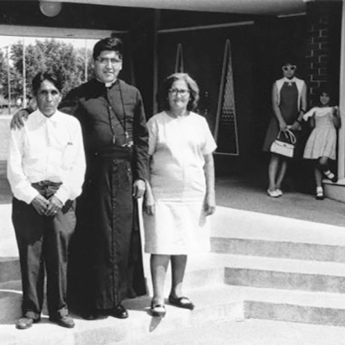 At the center stands Father Antonio Gonzales, a former migrant worker from Rio Grande City, Texas, who is visiting his parents in Wautoma. In 1966, he led a march for better working conditions in Texas. He has his arm around his father who is standing to the left with his hands folded in front of him. To the right is Father Gonzalez's mother wearing a dress and glasses. In the background to the right is a group of girls and a young woman wearing dresses and sunglasses.            This photograph is a part of Wisconsin-native David Giffey's series 'Struggle for Justice,'' images from the migrant farm worker struggle including an independent organizing effort in Wisconsin and the nationwide grape boycott movement started by Cesar Chavez of United Farm Workers during the 1960s and 1970s.            El Padre González en Wautoma            En el centro está el Padre Antonio Gonzales, él anteriormente fue un trabajador emigrante de Rio Grande City, Texas, visitando a sus padres en Wautoma. En 1966, él guio una marcha para mejorar las condiciones laborales en Texas. Él tiene su brazo alrededor de su padre que está parado a su izquierda y con las manos dobladas enfrente de él. A la derecha del Padre González está su madre en un vestido y con anteojos. Al fondo y hacia la derecha está un grupo de niñas y una mujer joven en un vestido y con lentes de sol. Esta fotografía es parte de la serie 'Lucha por la Justicia' tomada por David Giffey, originario de Wisconsin, las imágenes muestran la lucha de los trabajadores agrícolas emigrantes incluyendo un esfuerzo independiente organizado en Wisconsin y el movimiento nacional del boicot de uvas empezado por Cesar Chávez de la unión de campesinos o United Farm Workers durante los años 1960 y 1970.